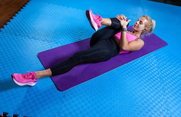 Slim fit blonde woman on mat doing warm-up exercises. Fit woman lies on yoga mat and practicing stretching legs and whole body. Healthy lifestyle, fitness concept