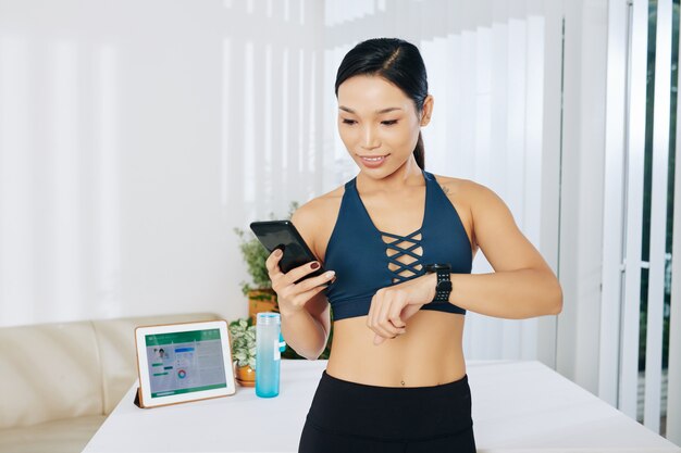 Slim fit jonge Aziatische vrouw die applicaties op haar smartwatch, smartphone en tablet synchroniseert nadat ze thuis heeft getraind