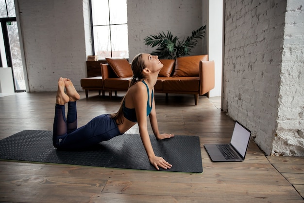 Slim fit woman practicing yoga and stretching body at home using laptop for online class or virtual tutorials. High quality photo