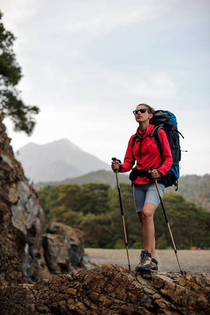 Slim fit woman in special wear standing with hiking backpack
and sticks on the rock