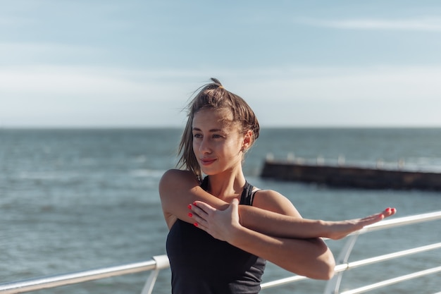 Slim fit woman in sportswear doing stretching hand exercise on the beach at bright sunny day