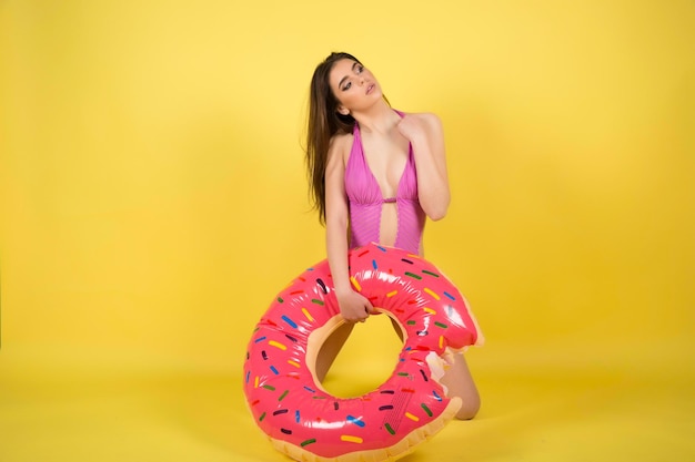 Slim girl in bikini with inflatable donut posing against yellow wall