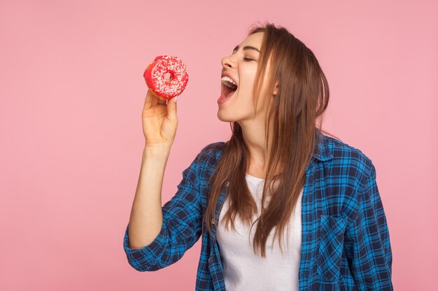 Slim girl in checkered shirt standing with eyes closed and holding her mouth open dreaming of biting tasty doughnut struggling temptation to eat sugary confectionery indoor studio shot isolated