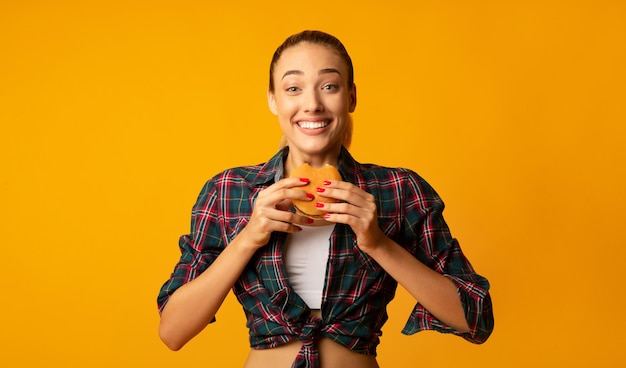 Фото Стройная девушка ест гамбургер на фоне желтой студии