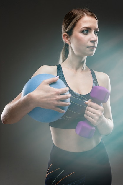 slim girl in sportswear holds a sports ball and a dumbbell on a black wall with lens flare effect