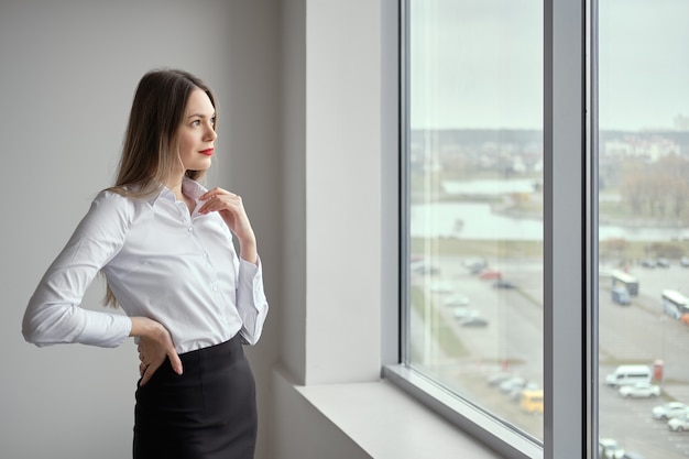 Худенькая девушка в белой рубашке и черной обтягивающей юбке позирует у окна
