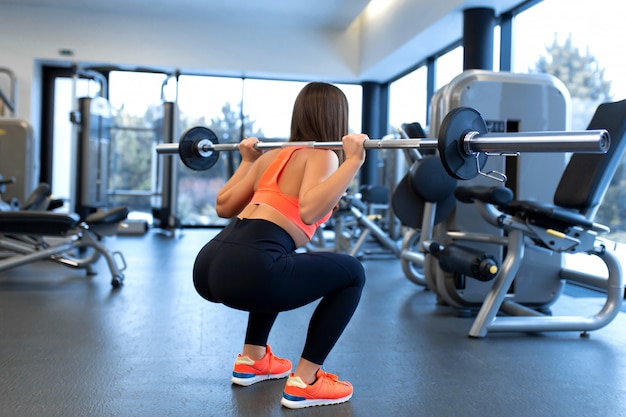 Slim handsome young woman in sportswear squats with a barbell on the shoulder in the gym