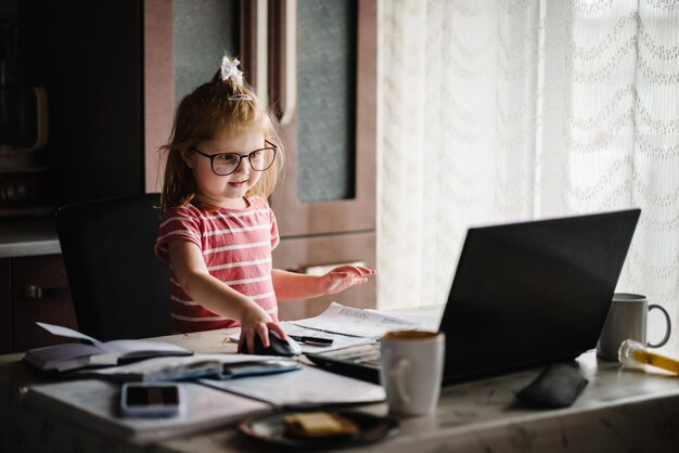 Foto slim peutermeisje dat een grote bril draagt tijdens het gebruik van haar laptop mooi meisje zit op de werkplek in het kantoor aan huis