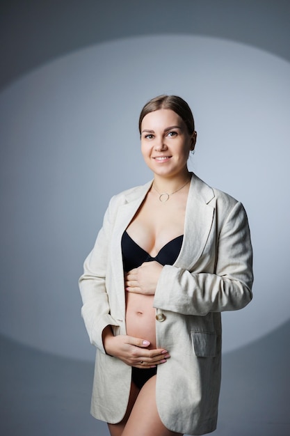 A slim pregnant woman in a long jacket and underwear stands in the studio on a white background holding her hand on her stomach Women's underwear for pregnant women
