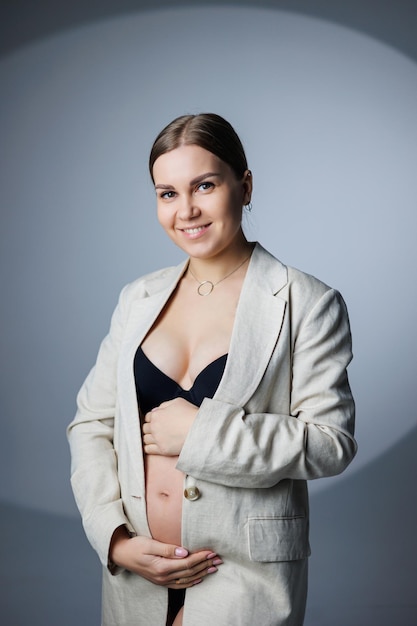 Стройная беременная женщина в длинной куртке и нижнем белье стоит в студии на белом фоне, держа руку на животе Женское нижнее белье для беременных