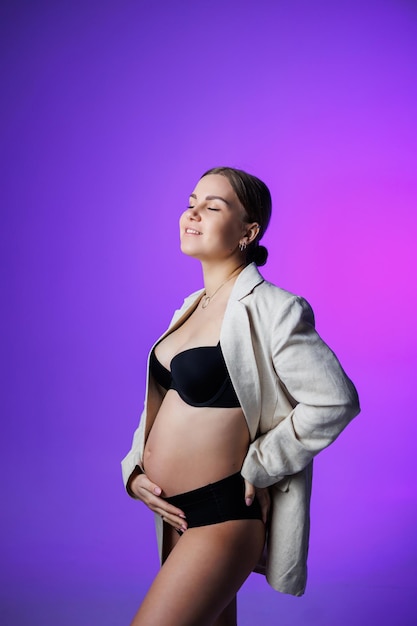 A slim pregnant woman in a long jacket and underwear stands in the studio on a white background holding her hand on her stomach Women's underwear for pregnant women