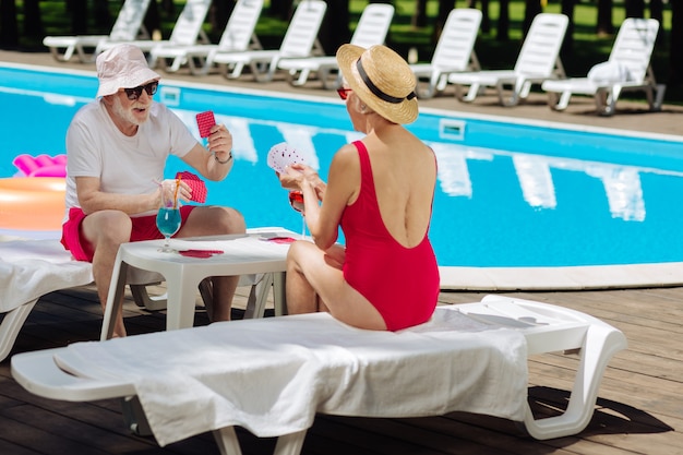 Slim retired woman wearing red swimming suit playing cards with her funny husband