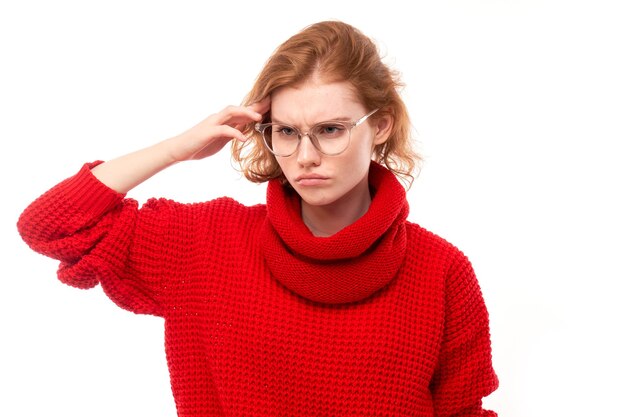 Slim roodharig meisje in het rood en een bril die haar hoofd aanraakt, denkt dat twijfels een besluit nemen geïsoleerd op een witte studioachtergrond