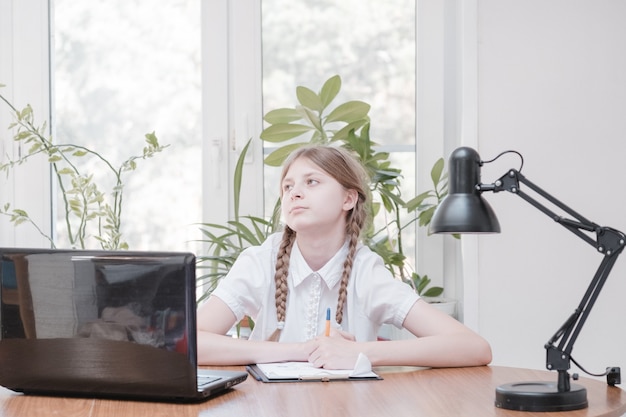 Slim schoolmeisje doet huiswerk zittend aan haar bureau, schrijft met een pen in haar schrijfnotities en gebruikt een laptop. Ze leert online taal, doet thuis schooltaken, luistert naar een lezing