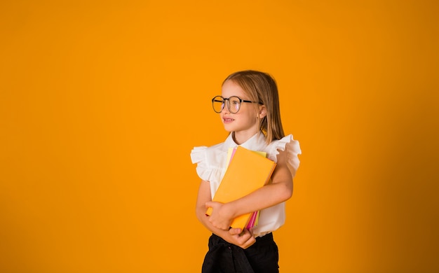 Slim schoolmeisje in een uniform met bril houdt boeken vast op een gele achtergrond met een kopie van de ruimte