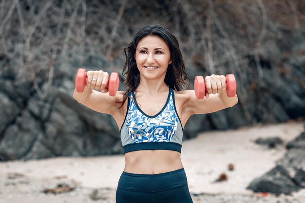 A slim smiling woman holds bumd-bells in front of her, doing sports at the beach