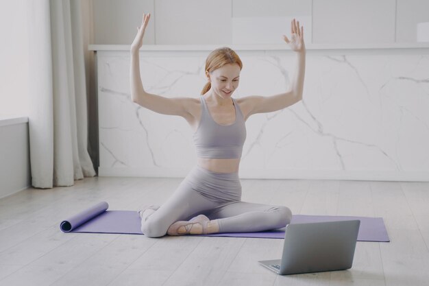 Slim sporty girl stretching on mat online remote fitness\
classes student watching video on laptop