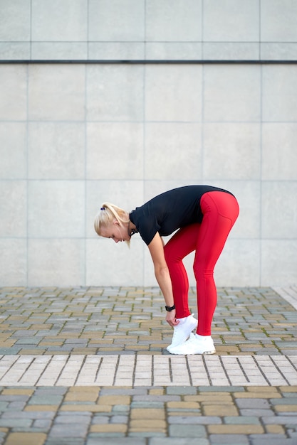 Slim sporty woman wearing stylish black sportswear, red leggings and trendy white sneakers lacing up sneakers