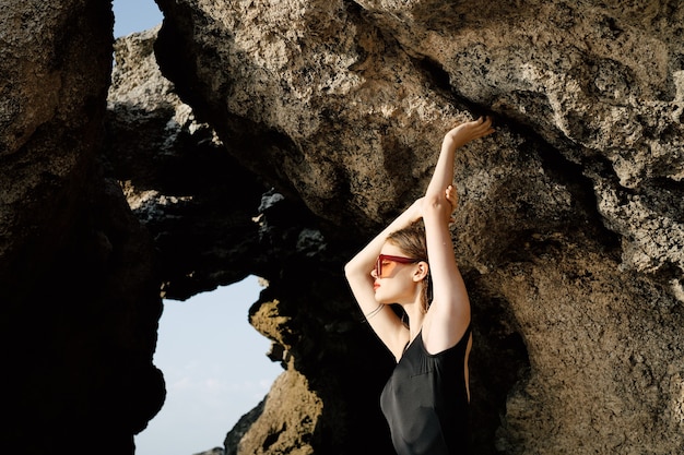 Slim woman in black swimsuit posing by the ocean against the backdrop of rocks fashion style