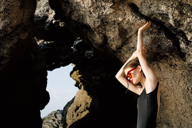 Slim woman in black swimsuit posing by the ocean against the backdrop of rocks fashion style