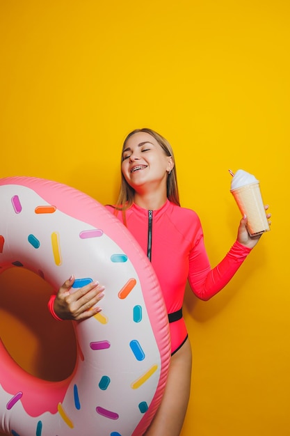 A slim woman in a bright pink swimsuit on a yellow background She is holding an inflatable pool donut Beach fashion