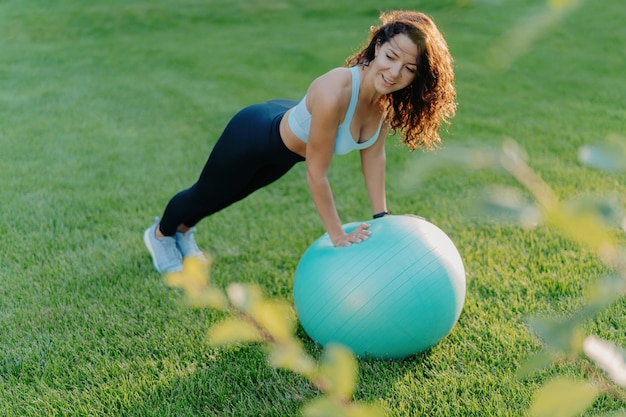 Slim woman enjoys gymnastic exercises on green lawn with fit ball wearing active wear Embraces healthy active lifestyle Fitness concept