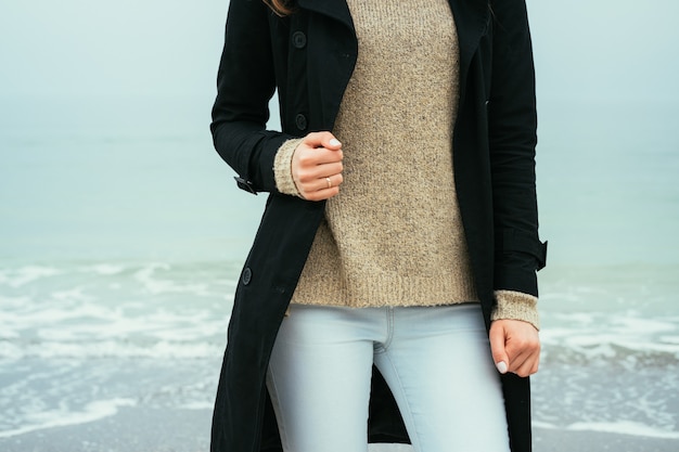 Slim woman in jeans, a sweater and a black coat against the sea on a cloudy day