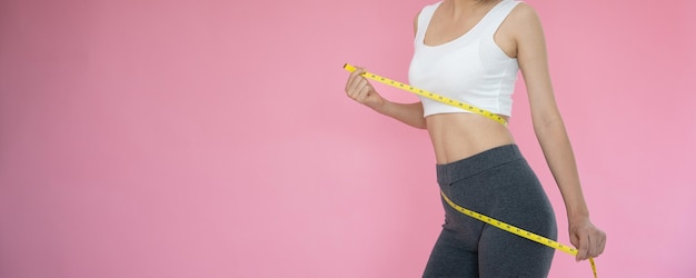 운동복을 입은 날씬한 여성은 분홍색 배경 다이어트 여성에 대한 줄자를 사용하여 허리를 측정하고 체중 감량 목표를 달성합니다.