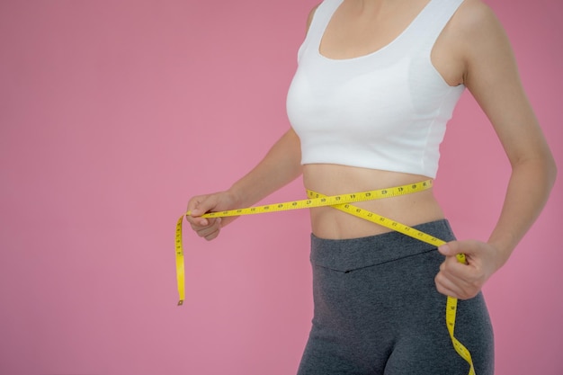 Slim woman in sportswear measures her waist using tape measure on pink background diet woman and lose weight plan