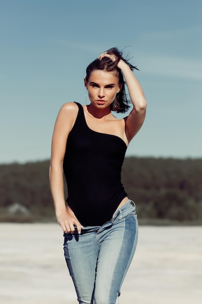 Slim woman in trendy bodysuit and jeans touching body while resting on beach on sunny and windy day