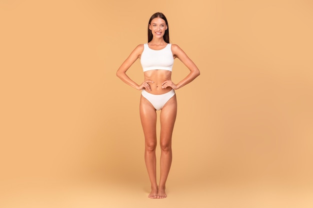 Foto donna snella in biancheria intima bianca con perfetta forma del corpo e pancia piatta in posa su sfondo beige per tutta la lunghezza