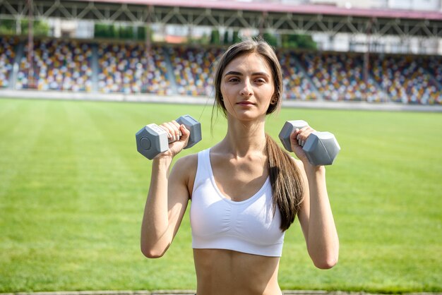 屋外でポーズをとるダンベルを持つスリムな女性。彼女は運動をしていて、スポーツスーツを着ています