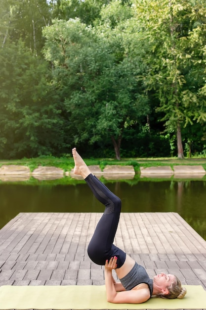 A slim woman on a wooden platform by a pond in a park in summer does yoga