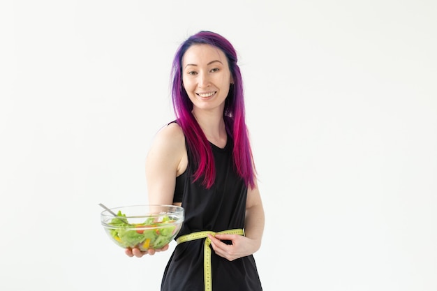 Slim young asian girl with colored hair is measured hanging a measuring tape and holding a light vegetable salad in her hands posing on a white wall. Healthy eating concept. Copyspace.