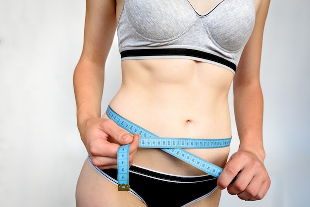 Slim young girl measuring her waist with blue tape measure on gray background. Slim and fitness concept.