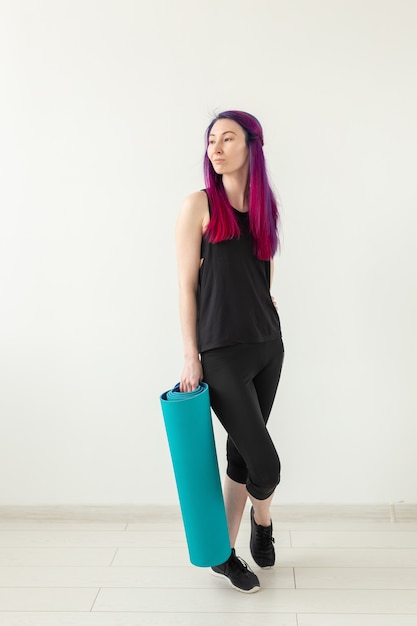 Slim young mixed race girl hipster with colored hair folds gymnastic mat after yoga in the gym. Healthy lifestyle and fitness concept.