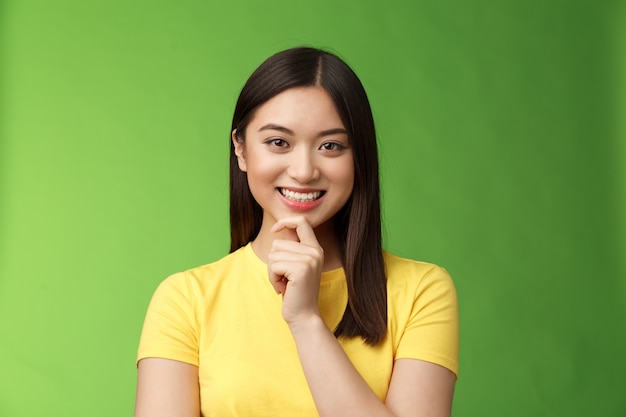 Slimme assertieve creatieve aziatische vrouwelijke student maakt interessante onderzoeksaannames, glimlacht geïntrigeerd en raakt kin attent, heeft een goed planidee, denkt na, overdenkt keuzes, groene achtergrond.