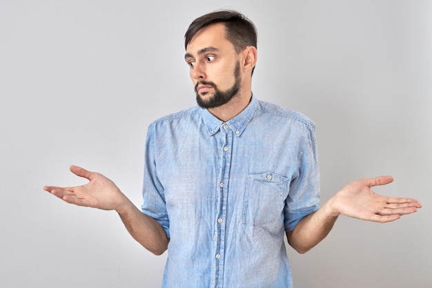 Slimme brunette man in casual schouderophalende handen denkt dat twijfels een besluit nemen geïsoleerd op blauwe studio achtergrond