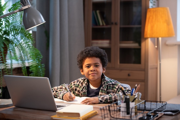 Slimme en ijverige Afrikaanse schooljongen die aantekeningen maakt in het schrijfboek en naar je kijkt terwijl hij aan de tafel zit voor de laptop in de woonkamer