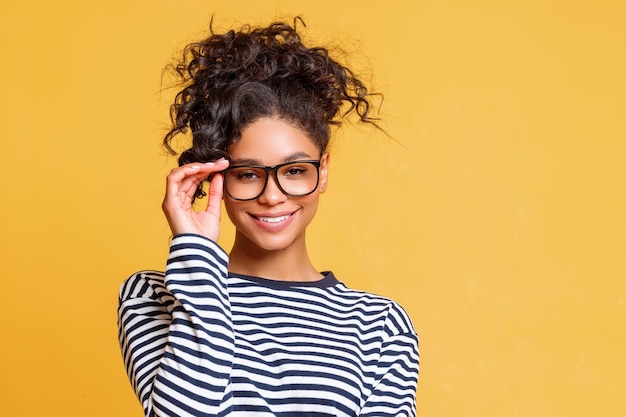 Slimme etnische vrouw in gestreept kledingstuk dat lacht voor de camera en een trendy bril aanpast tegen gele achtergrond