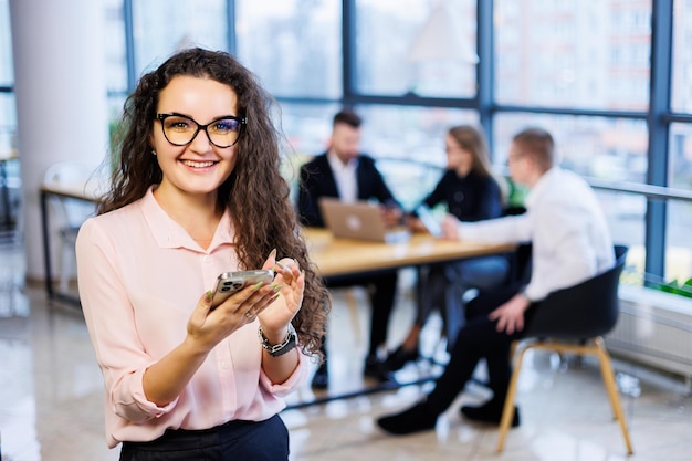 Slimme gelukkige jonge zakenvrouw, in stijlvolle kleding en bril, werkt op kantoor, praat met een klant op een smartphone, gebaren, glimlacht. op de achtergrond zijn medewerkers aan het werk. selectieve focus