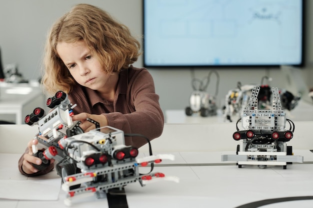 Slimme jeugdige schooljongen die bij het bureau zit en speelgoedrobot bouwt