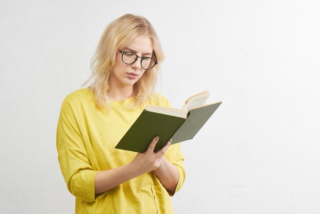 Slimme jonge blonde met bril leest een boek met een serieus gezicht in gele kleren alleen op een witte studioachtergrond