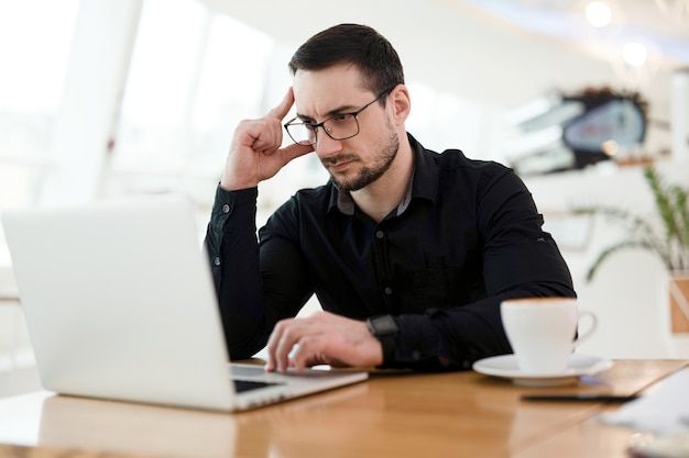 Slimme jonge professional denken over technische taak. Geconcentreerde man die zijn hoofd vasthoudt en laptop gebruikt. Werkconcept op afstand. Kopje lekkere cappuccino.