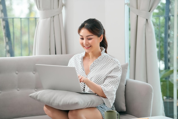 Slimme jonge vrouw met een bril die op de bank zit en freelance project doet op laptop in een gezellige woonkamer thuis