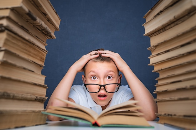 Slimme jongen in glazen gek van het lezen van boek zittend tussen twee stapels boeken