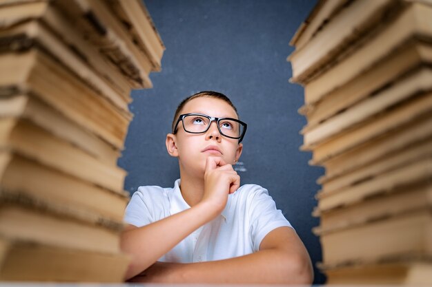 Slimme jongen in glazen zitten tussen twee stapels boeken en bedachtzaam opzoeken.