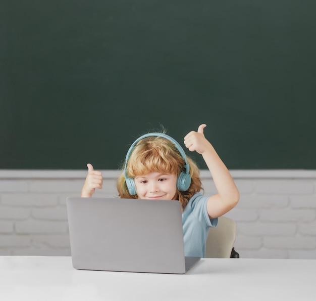 Slimme jongen met duimen omhoog in koptelefoon zit aan bureau online studeren op laptop op school klever kid wear