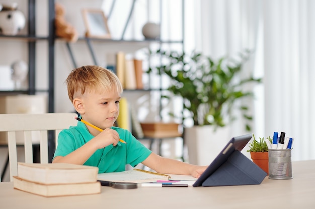 Slimme kleine jongen zit aan een bureau en kijkt naar onderwijsvideo's op tabletcomputer of volgt online les