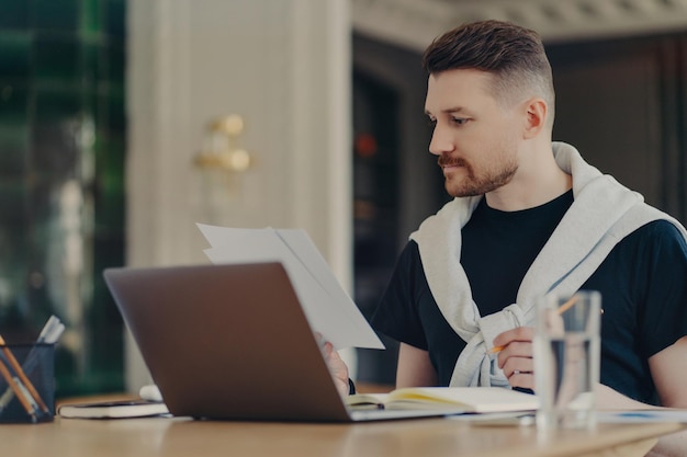 Slimme knappe man controleert informatie over papieren werkt op afstand zit op kantoor aan huis maakt gebruik van moderne laptopcomputer schrijft op in notebook gekleed in vrijetijdskleding Freelance werk en technologie